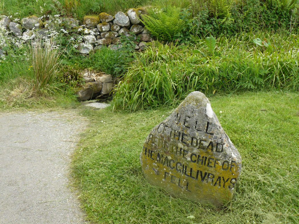 Grabstein auf einer Wiese neben einem Weg, im Hintergrund eine Mauer aus Findlingen